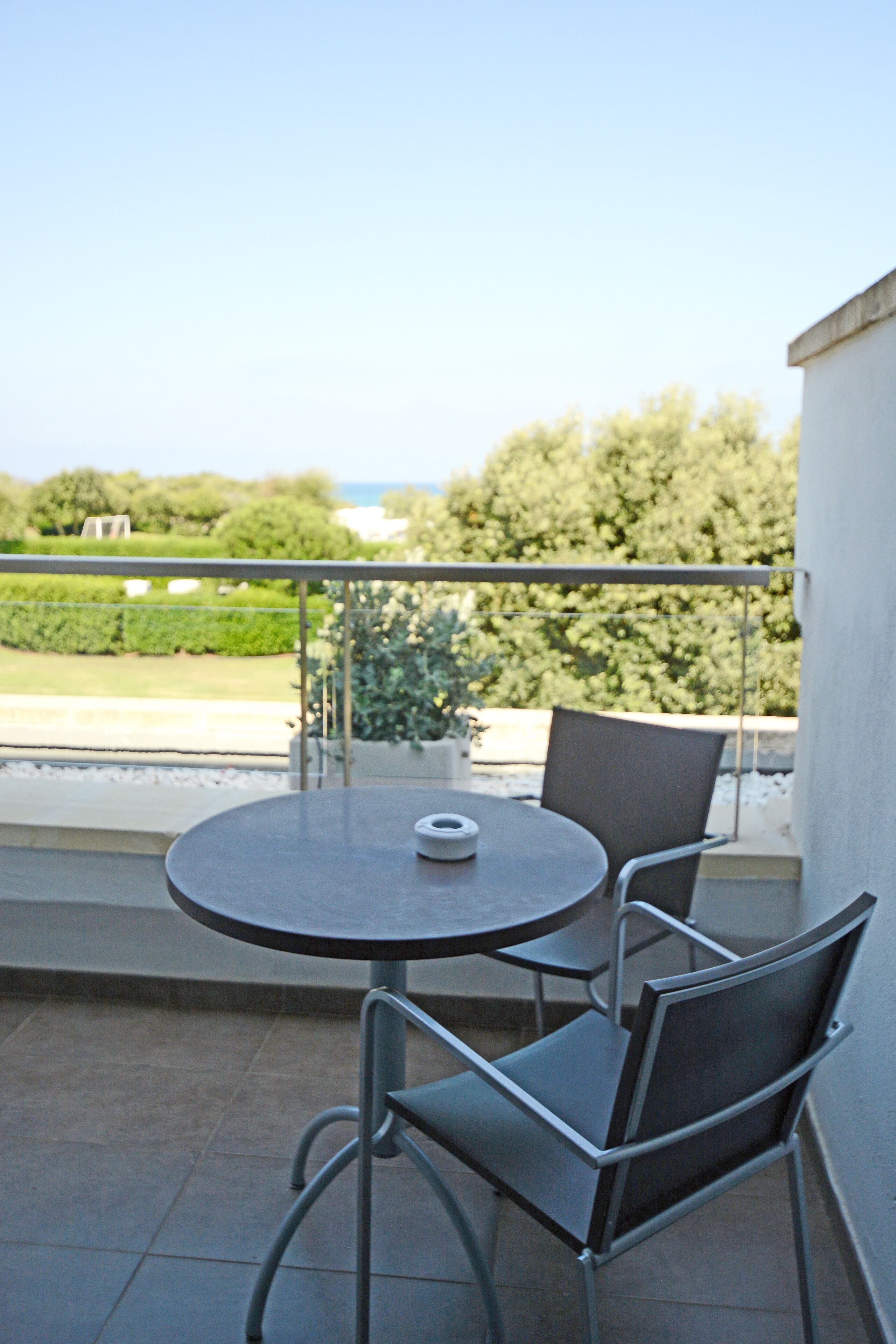 A balcony with a round table and two chairs, a potted plant, and a view of a garden and trees.