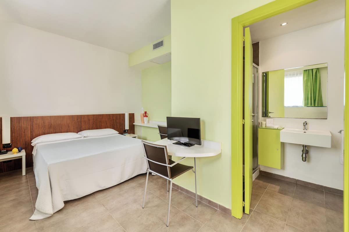 A hotel room with a bed, desk, chair, and a bathroom with a sink and mirror.