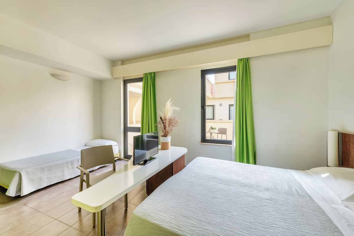 A hotel room with two single beds, a desk with a chair, a TV, and a window with green curtains.