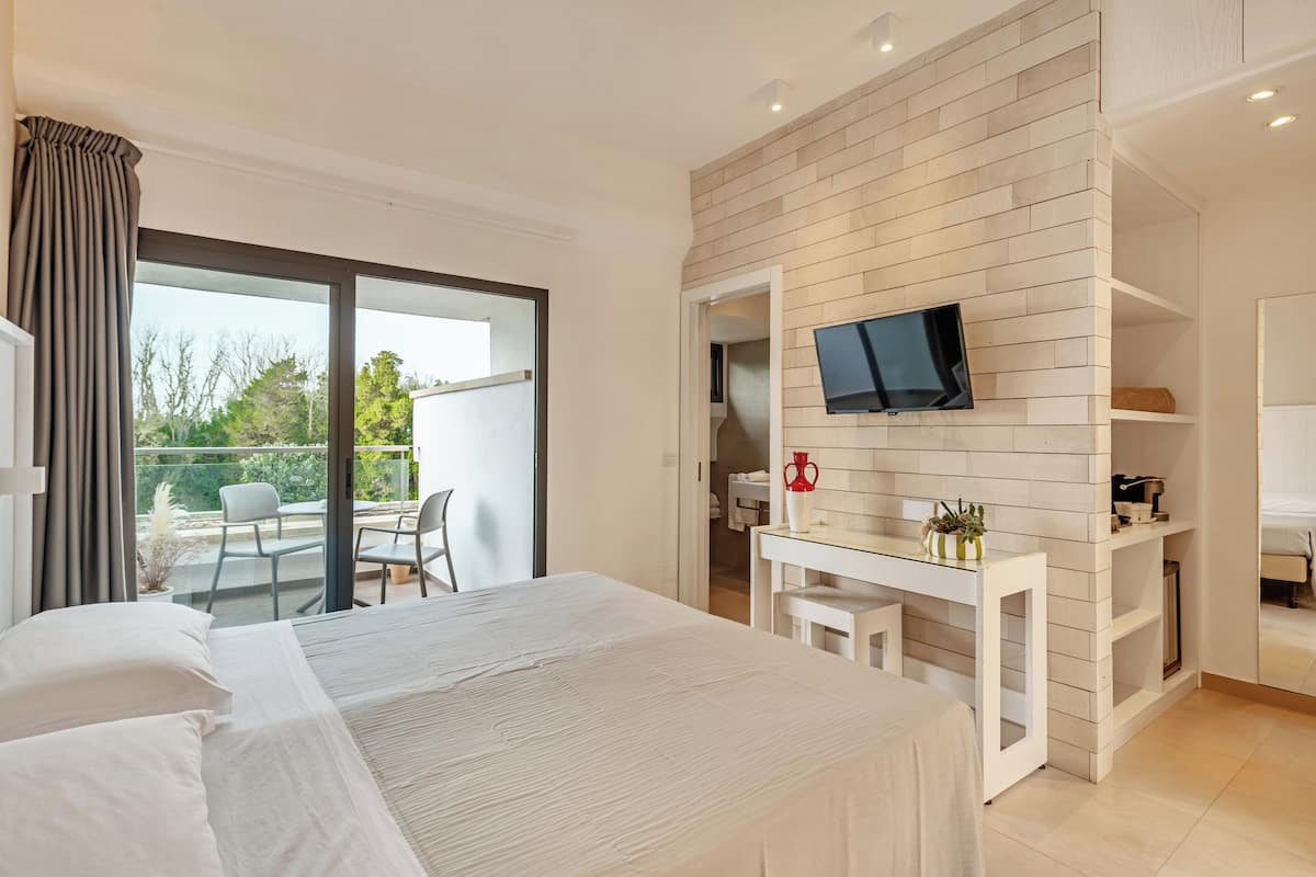 A modern hotel room with a large bed, a balcony with a table and chairs, a TV mounted on a wooden wall, and a small desk with a vase and plants.