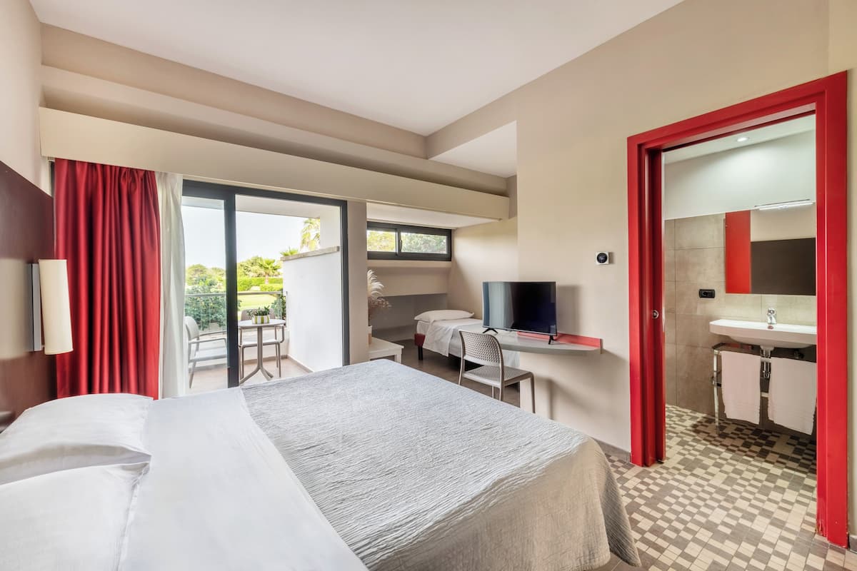 A hotel room with a bed, a desk, a chair, a TV, a bathroom with a sink and mirror, and a view of the outdoors.