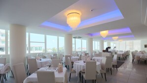 A spacious dining area with multiple round tables and chairs, large windows, and a modern chandelier. - Hotel King (Cervia)