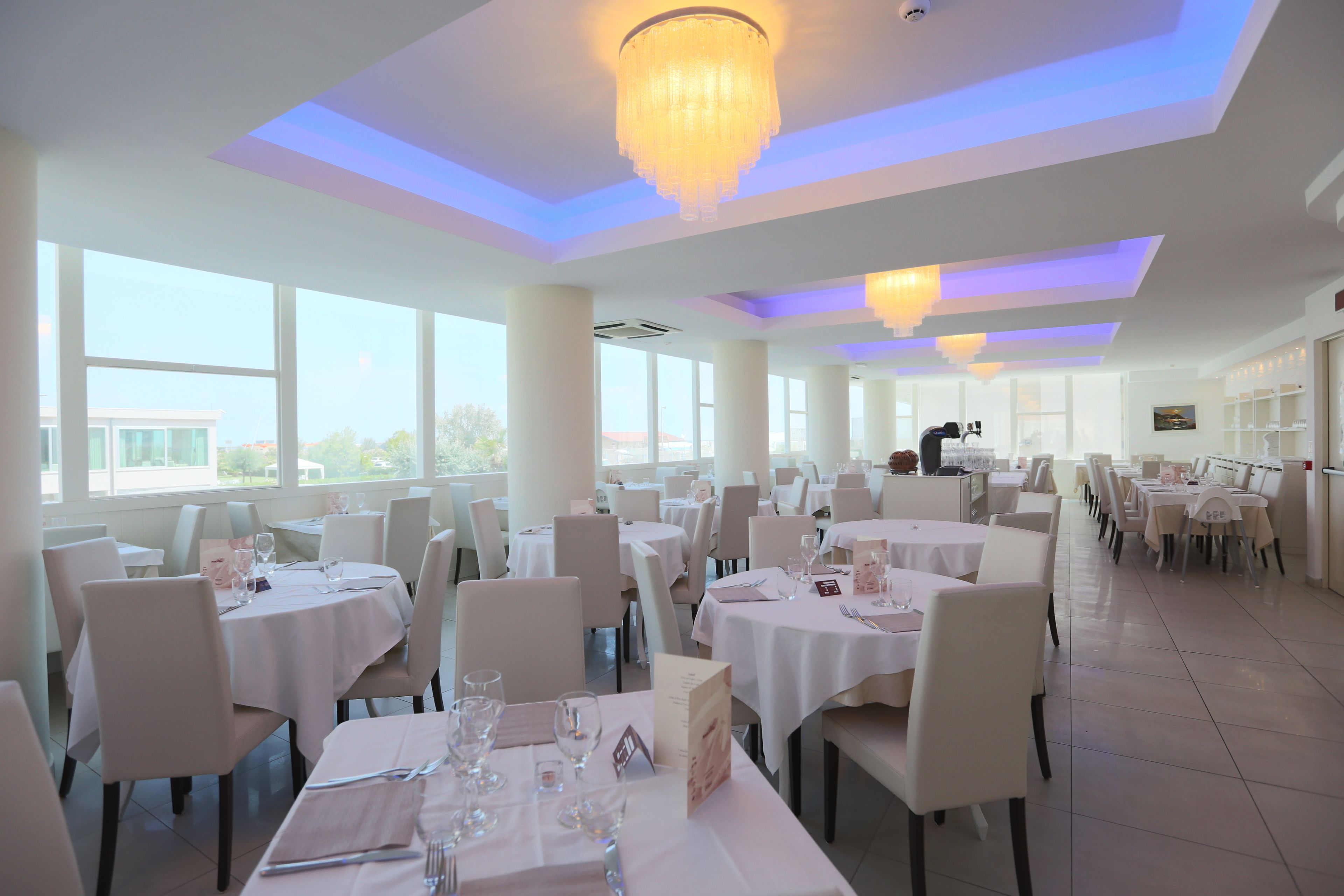 A spacious dining area with multiple tables and chairs, large windows, and a modern chandelier.