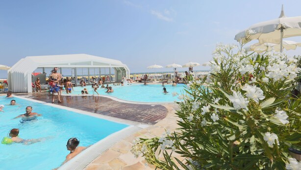 A pool area with people swimming and lounging, surrounded by white umbrellas and a covered seating area. - Hotel King (Cervia)