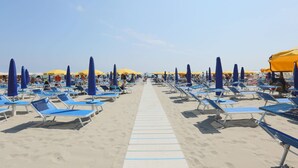 A beach with rows of blue and yellow sun loungers and umbrellas, a sandy walkway, and a clear blue sky. - Hotel King (Cervia)
