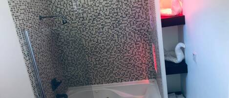 A bathroom with a mosaic-tiled shower enclosure, a white bathtub, and a shelf with towels.