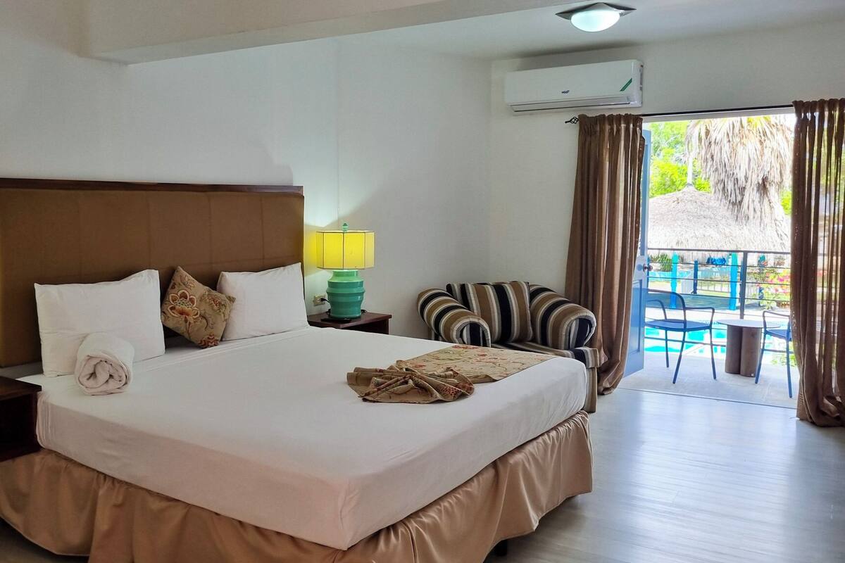 Une chambre d’hôtel avec un grand lit, une table de chevet avec une lampe, une chaise et une vue sur l’espace extérieur abritant une piscine.
