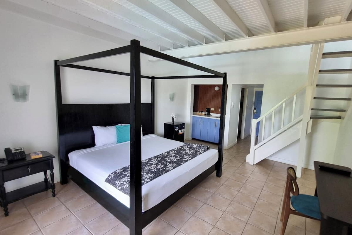Un lit à baldaquin avec une couvre-lit noire et blanche, une petite table de chevet avec un téléphone et une chaise. On aperçoit, au fond, une kitchenette avec des placards bleus et une cuisinière.