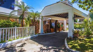 A covered entrance to a building with a white fence, palm trees, and a paved driveway.