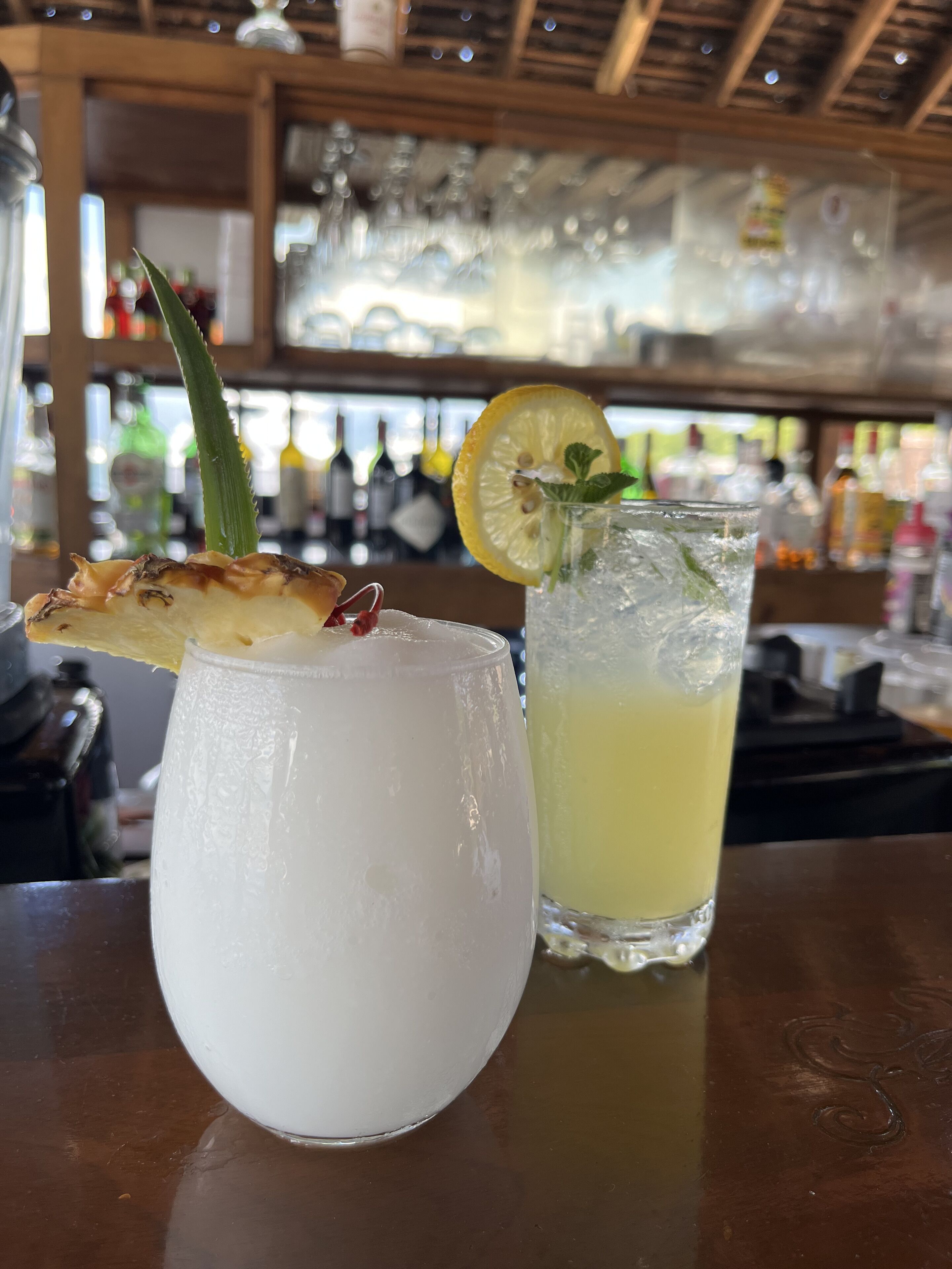 A bar setting with two drinks, one with a pineapple garnish and mint, and the other with a lemon slice and mint.