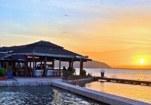 A waterfront restaurant with outdoor seating, a covered patio, and a view of the sunset over the water. - Grand Hotel Excelsior Port Royal (Kingston)