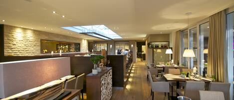 Un hall d’hôtel moderne doté d’un bar, de différents espaces aménagés pour s’asseoir et d’une verrière.