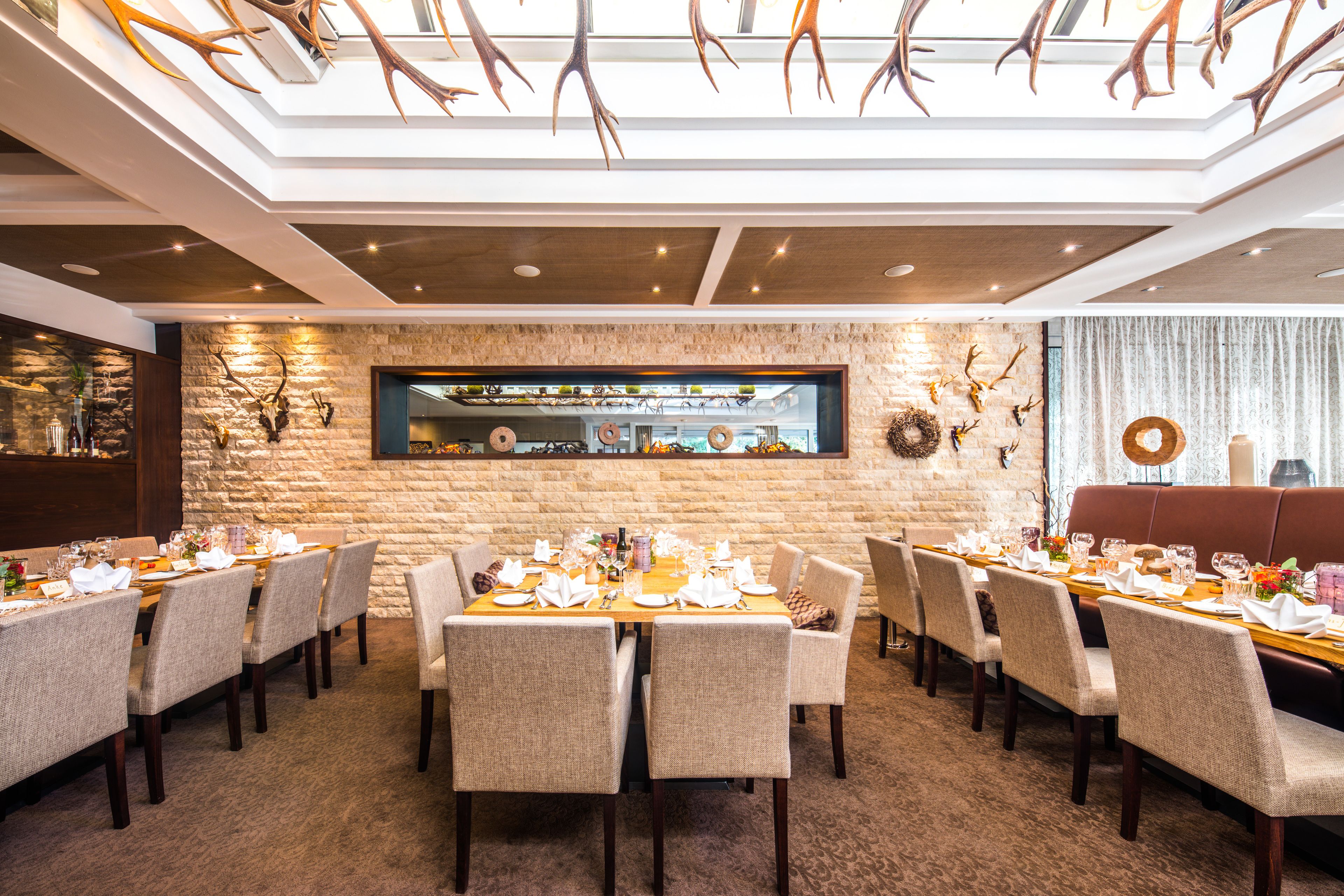 Comedor con techo alto, pared de piedra y decoración de cornamentas. Las mesas están dispuestas con manteles blancos y sillas.