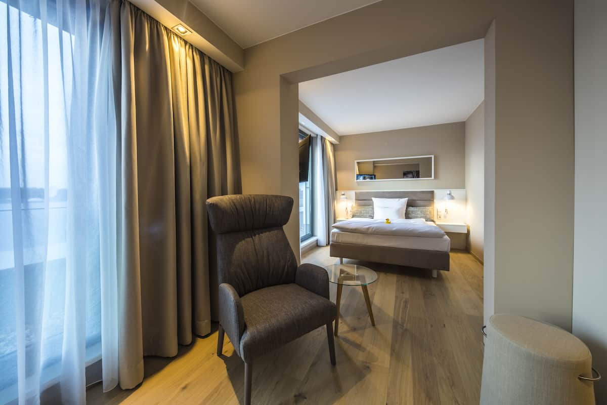 Een moderne hotelkamer met een groot raam, een comfortabele fauteuil, een glazen salontafel, een bed met een witte dekbedovertrek en een nachtkastje met een lamp.