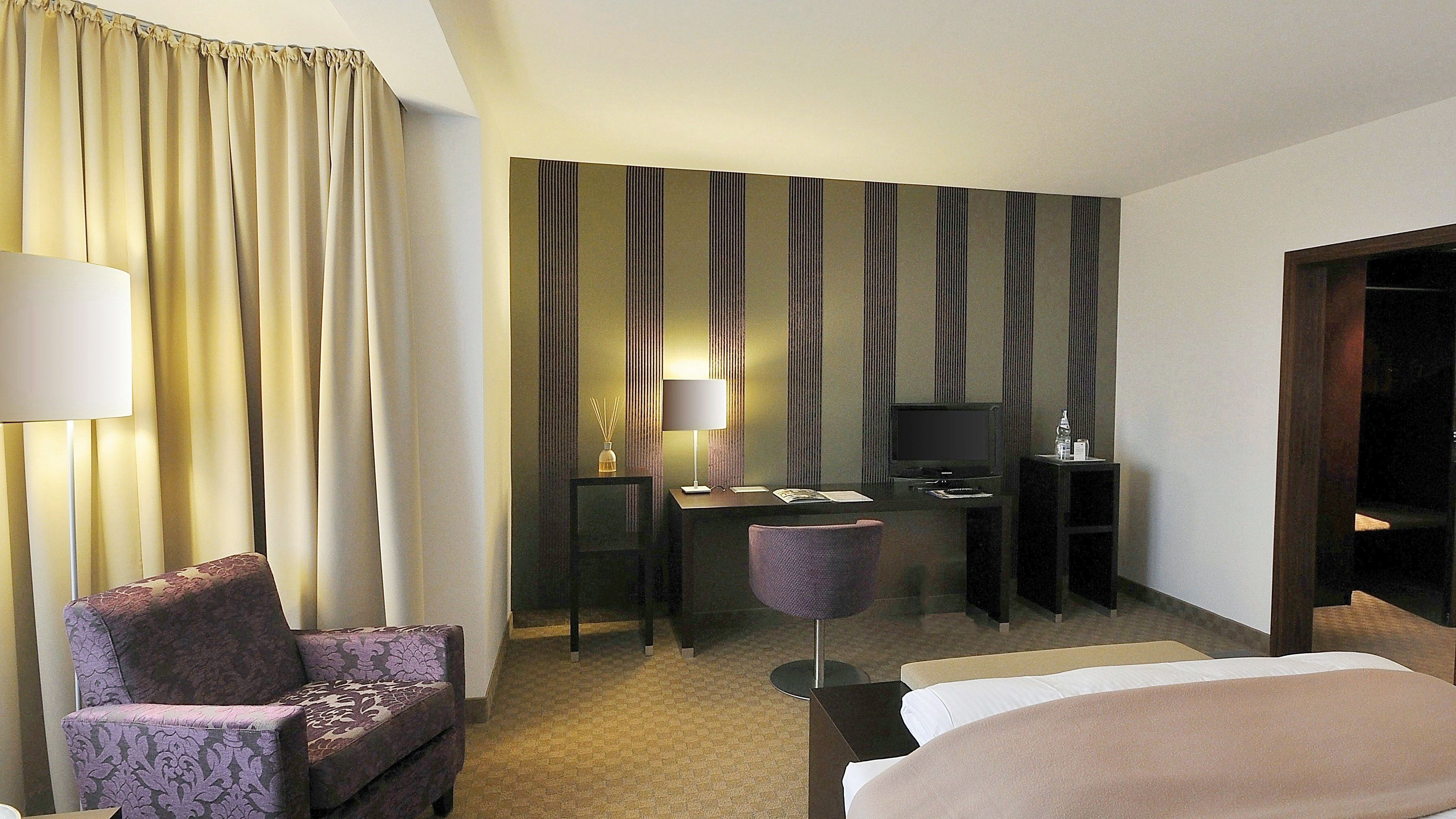 Quarto de hotel com cama, escrivaninha com computador, cadeira e luminária.
