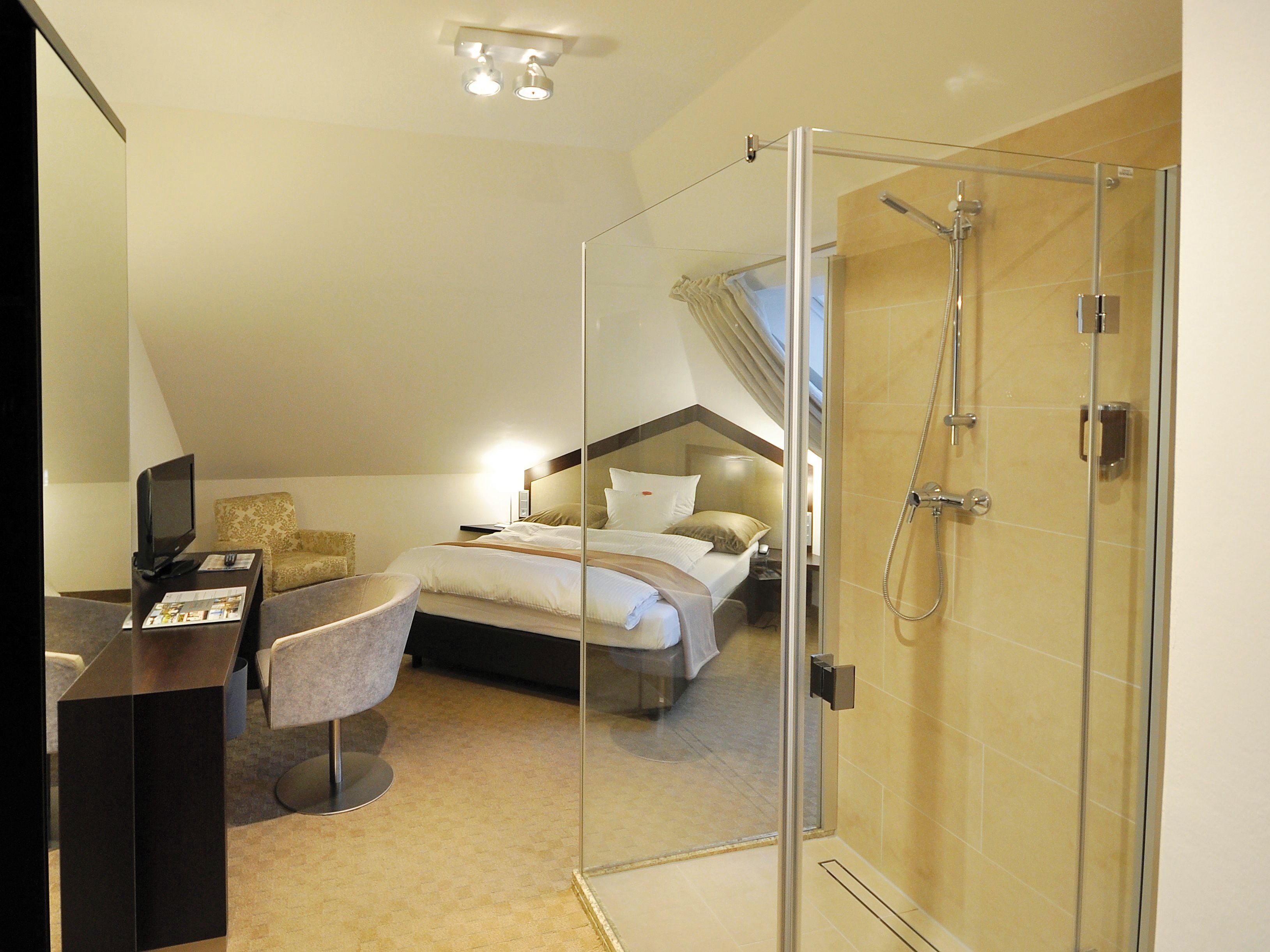 A modern hotel room with a glass-enclosed shower, a bed with a headboard, a desk with a computer, and a seating area.