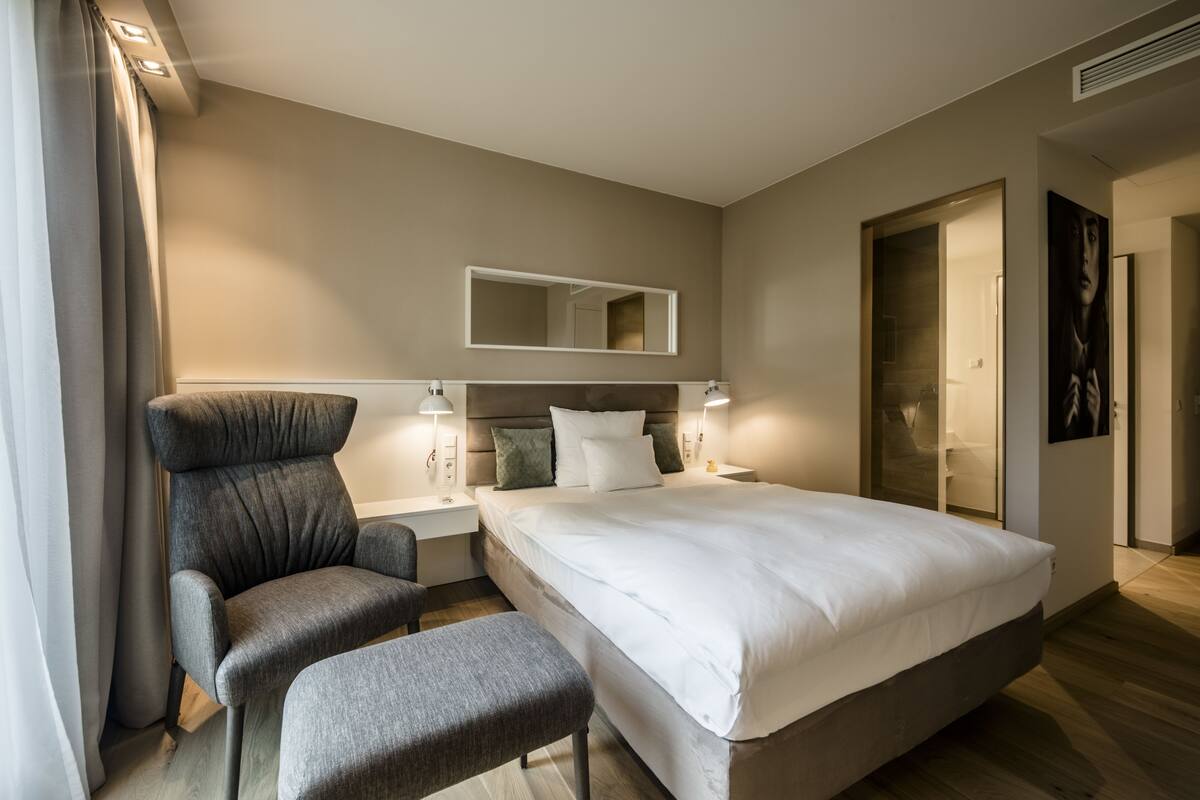 Een moderne hotelkamer met een groot bed, een grijze fauteuil, een klein tafeltje en een bedlampje.