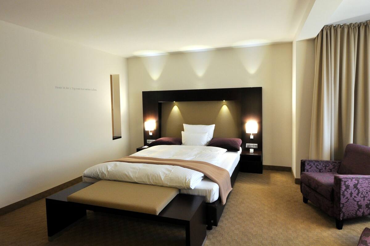 Een hotelkamer met een groot bed, een stoel, een klein tafeltje en een wandlamp.