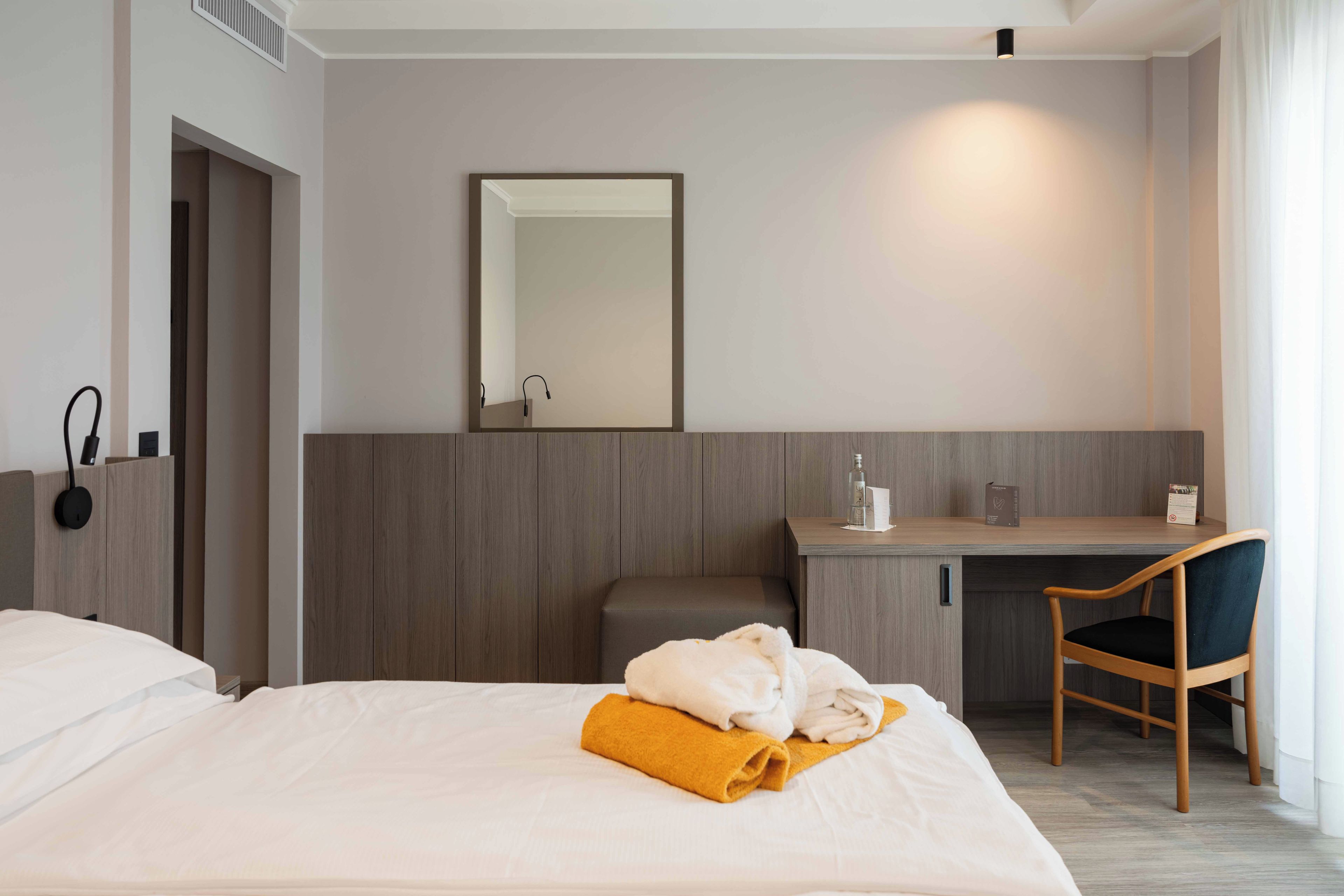 A hotel room with a bed, a desk, a chair, a mirror, and a nightstand.