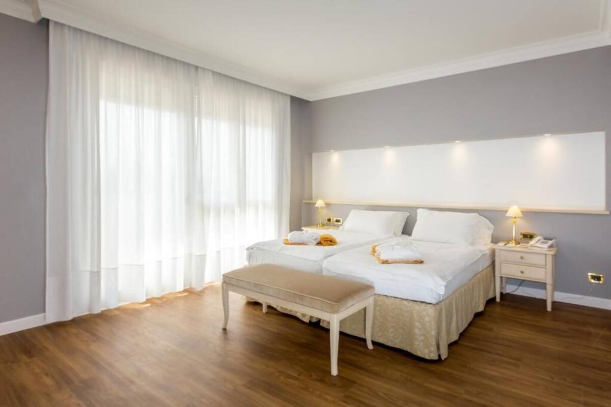 A bedroom with two beds, a bench, bedside tables, and a large window with curtains.