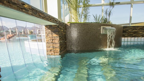 An indoor swimming pool with clear water, a waterfall feature, and a stone wall. - Hotel Terme Leonardo (Abano Terme)