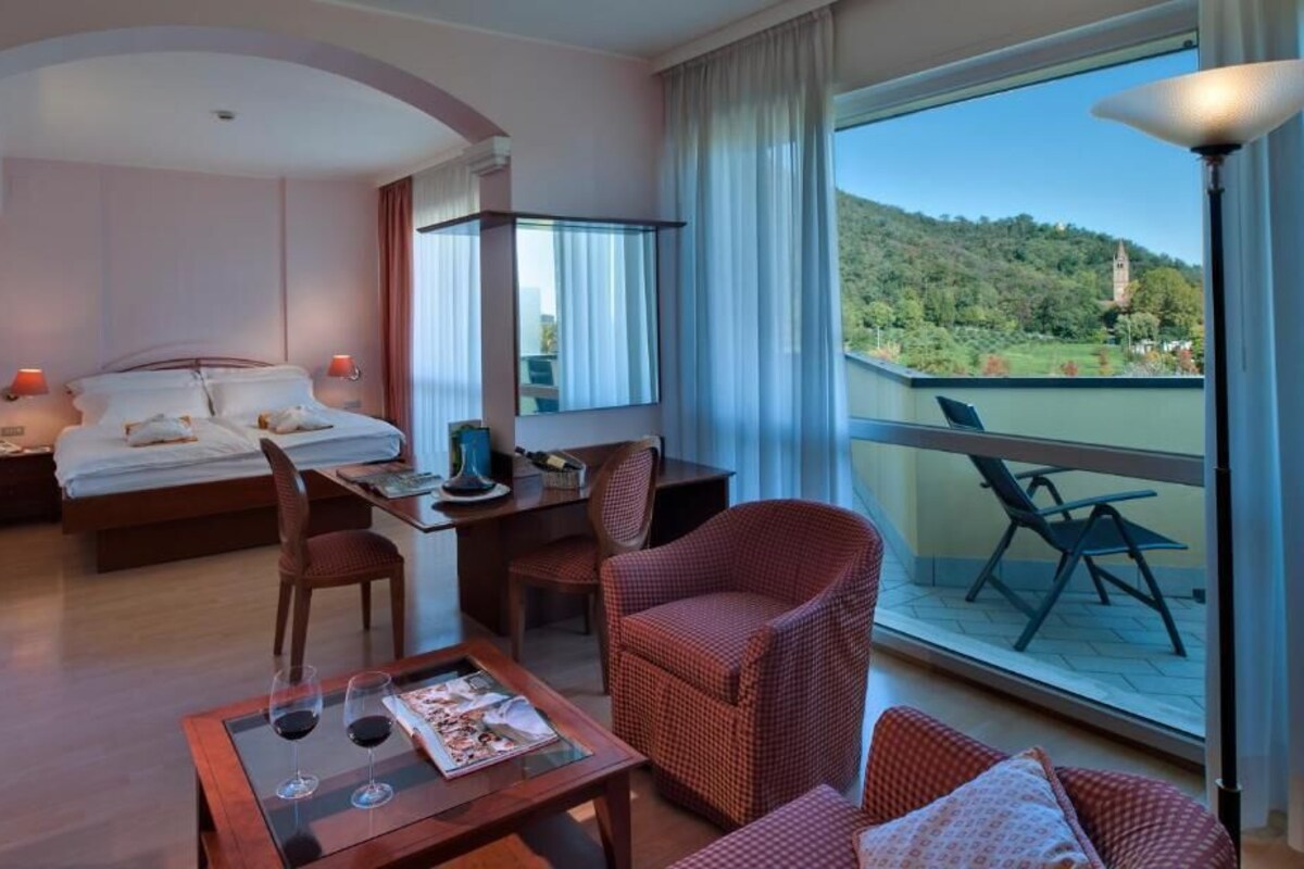 A hotel room with a bed, a desk, a dining table, and a balcony with a view of a hill and a tower.