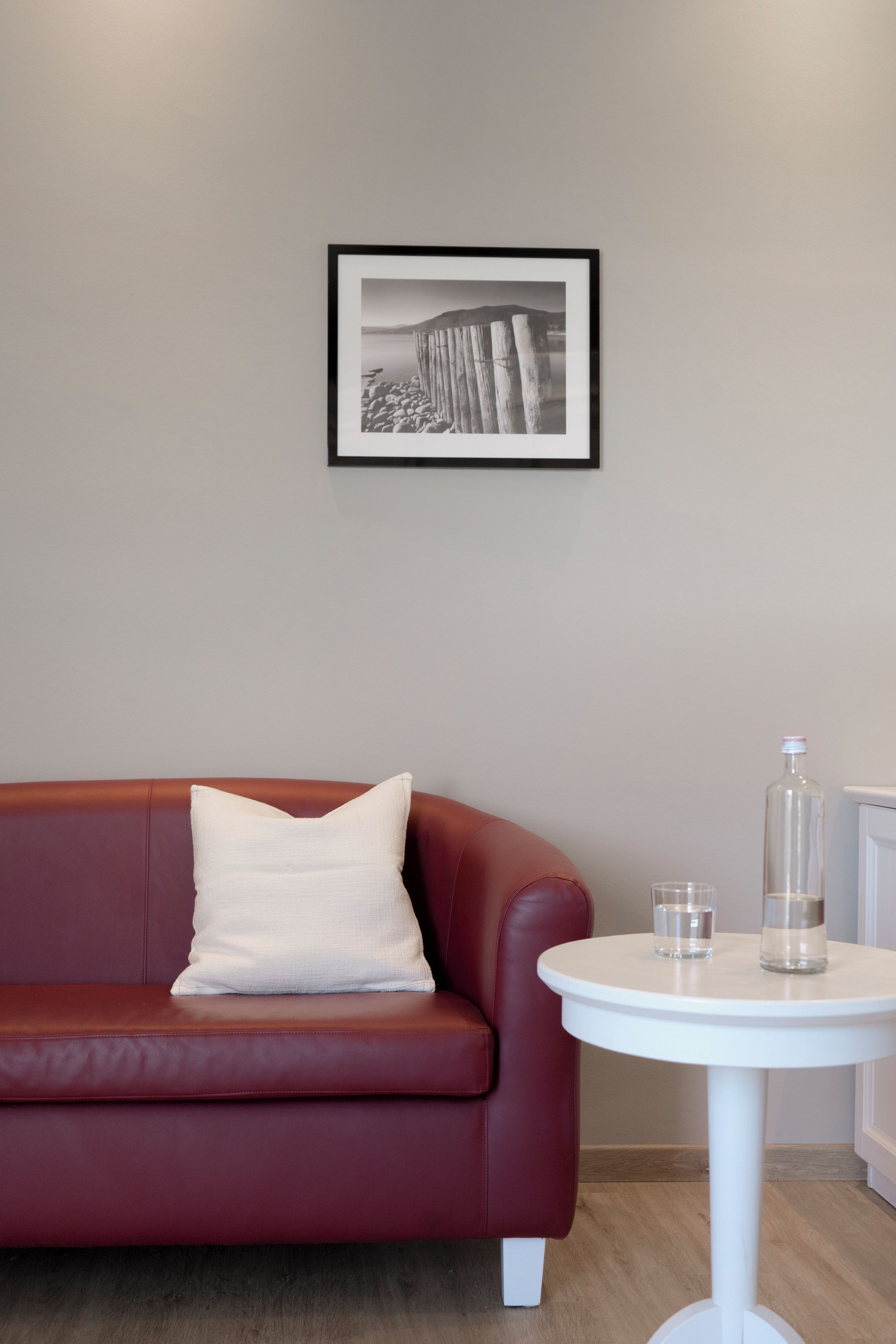 Un sofá de cuero rojo con un almohadón blanco, una fotografía en blanco y negro enmarcada en la pared, y una mesita blanca con dos vasos y una botella.