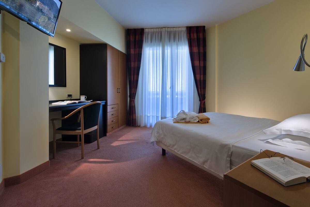 A hotel room with a bed, a desk, a chair, a TV, and a window with curtains.