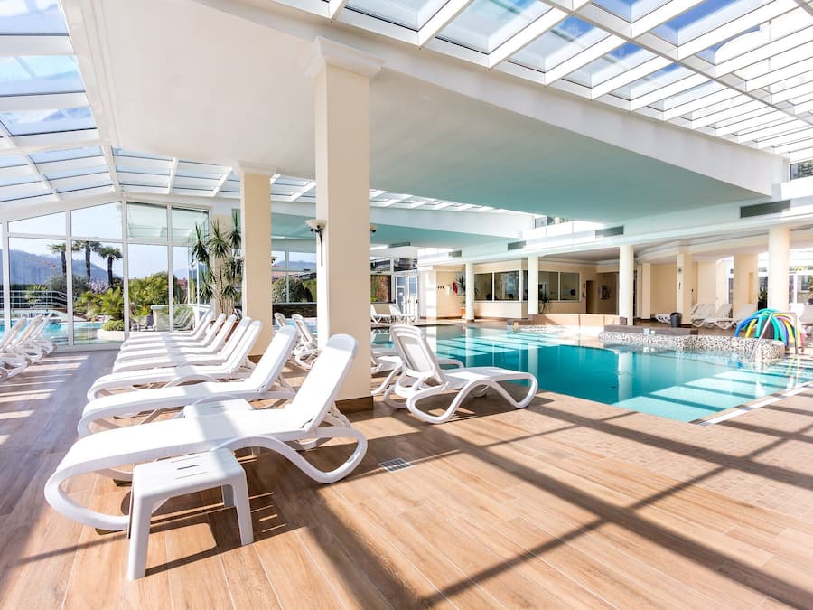 A sunlit indoor swimming pool with lounge chairs and a glass ceiling.