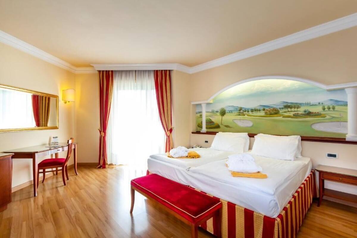 A hotel room with two beds, a desk, a chair, and a large painting of a golf course.