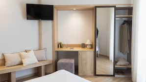Habitación de hotel con cama, escritorio con silla, televisor montado en la pared, armario con ropa colgada y un taburete pequeño.