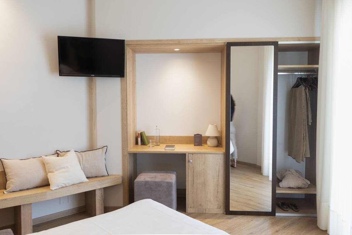 A hotel room with a bed, a desk with a chair, a TV mounted on the wall, a closet with hanging clothes, and a small stool.