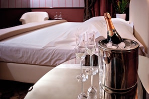 Una cama con sábanas blancas, una mesita de noche con una botella de champán y copas, y una planta en maceta al fondo.