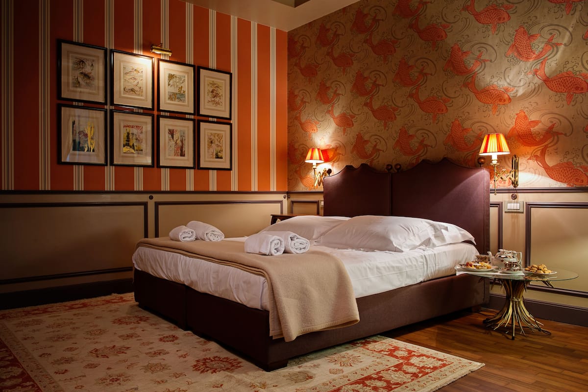 A neatly made bed with white linens and a beige throw blanket, a small round table with a lamp, and a wall with striped wallpaper and framed pictures.