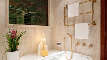Een badkamer met een marmeren wand, een bad, een gouden douchesysteem en een potplant met roze bloemen.