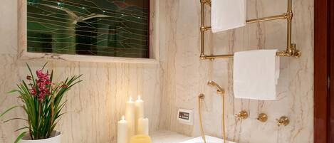 A bathroom with a marble wall, a bathtub, a gold shower system, and a potted plant with pink flowers.