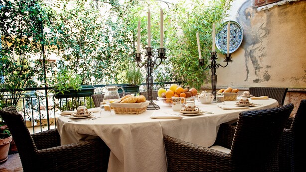 Un área de comedor al aire libre con sillas de mimbre y una mesa dispuesta para una comida, rodeada de follaje y un muro rústico.