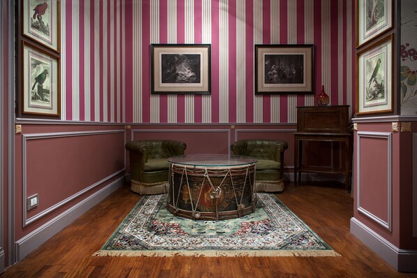 Una habitación con un escritorio de tambor, sillones verdes, un tapete estampado y cuadros en la pared.