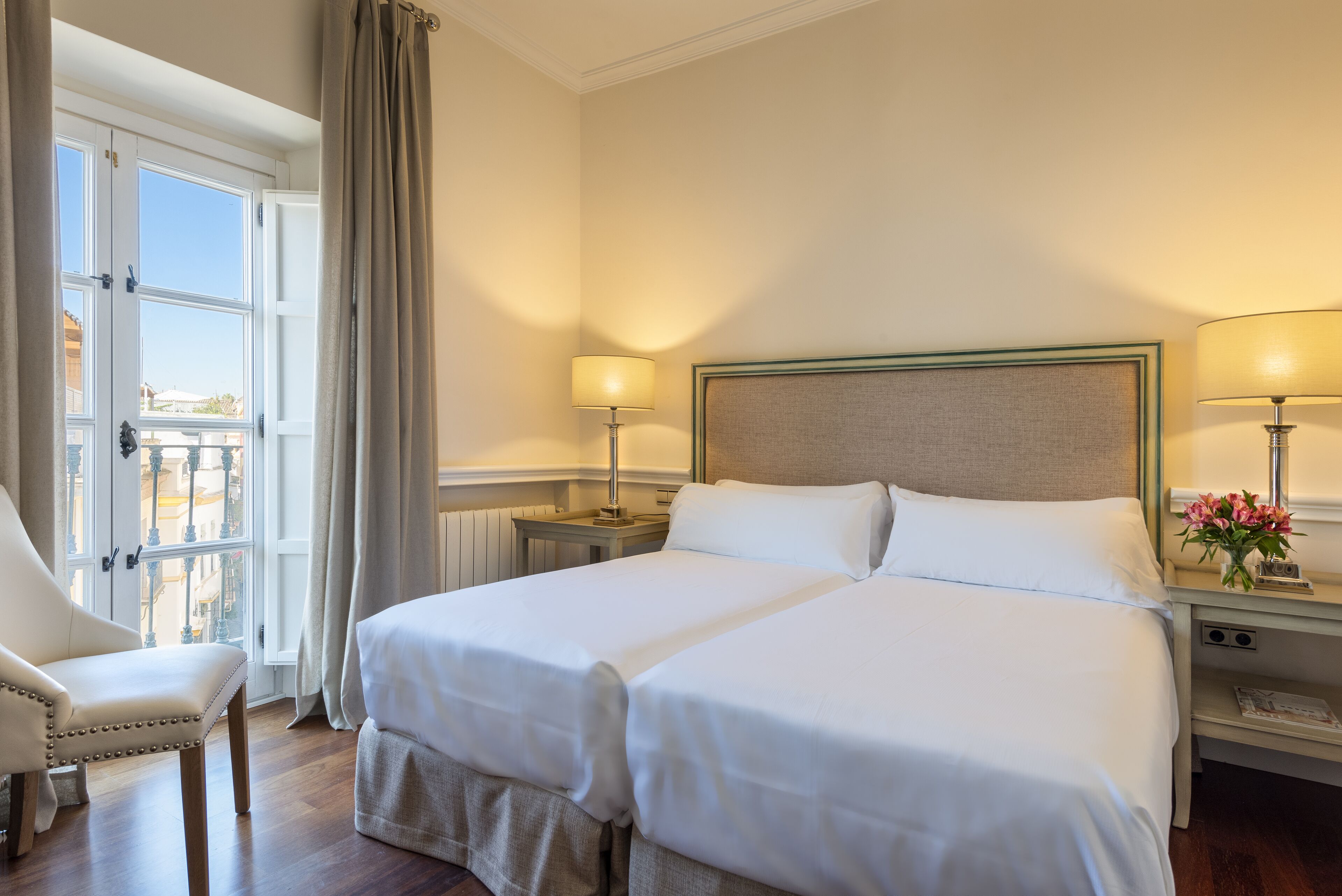 A double bed with white linens, two bedside tables with lamps, a chair, and a view of the outside through a window with curtains.