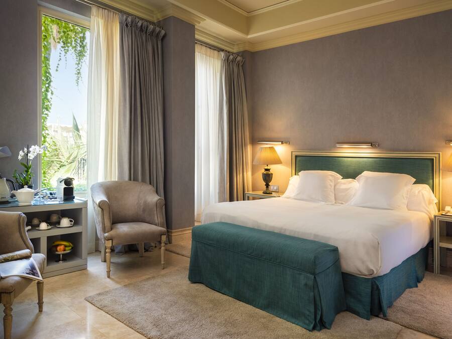 Habitación de hotel con una cama grande, dos sillas, un escritorio con lámpara y vistas a zonas verdes.