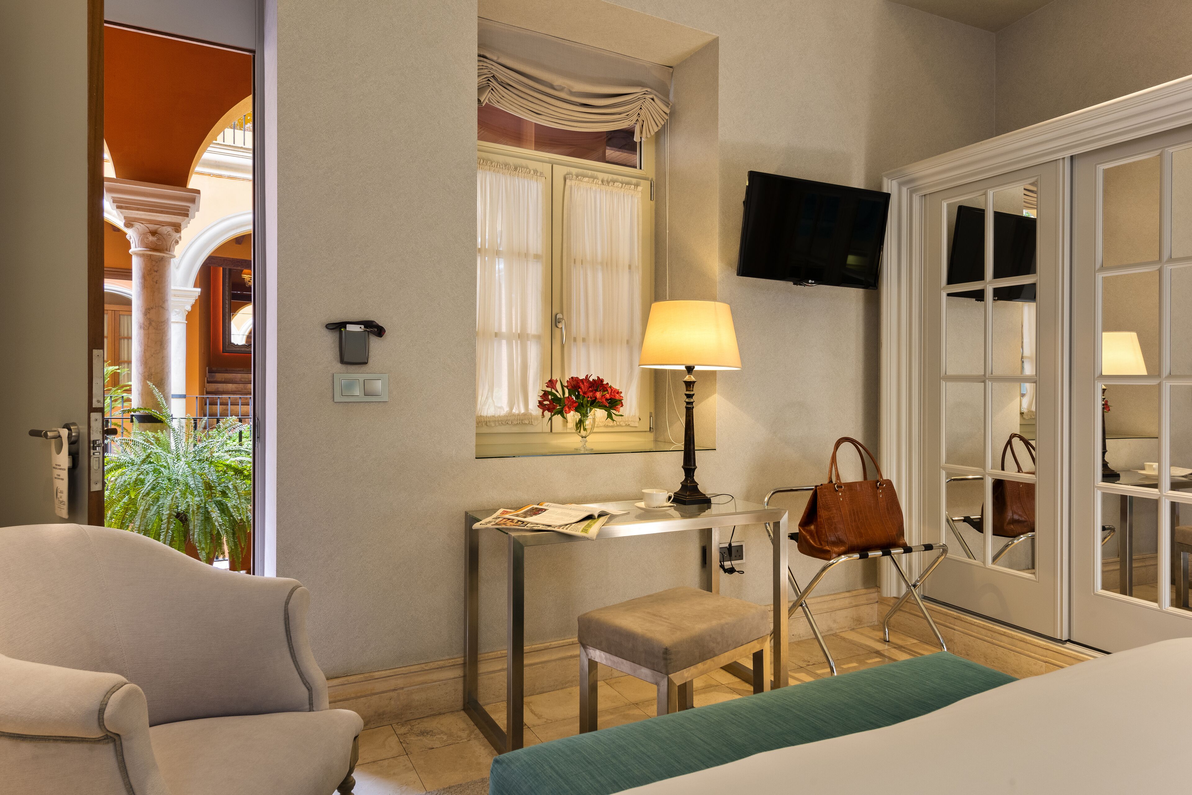 A hotel room with a bed, a desk with a lamp, a chair, a TV mounted on the wall, and a window with curtains.