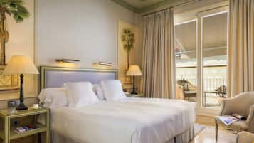 Habitación de hotel con una cama grande, dos mesitas de noche con lámparas, una silla, una pequeña mesa con una revista y vistas a un balcón con cortinas.