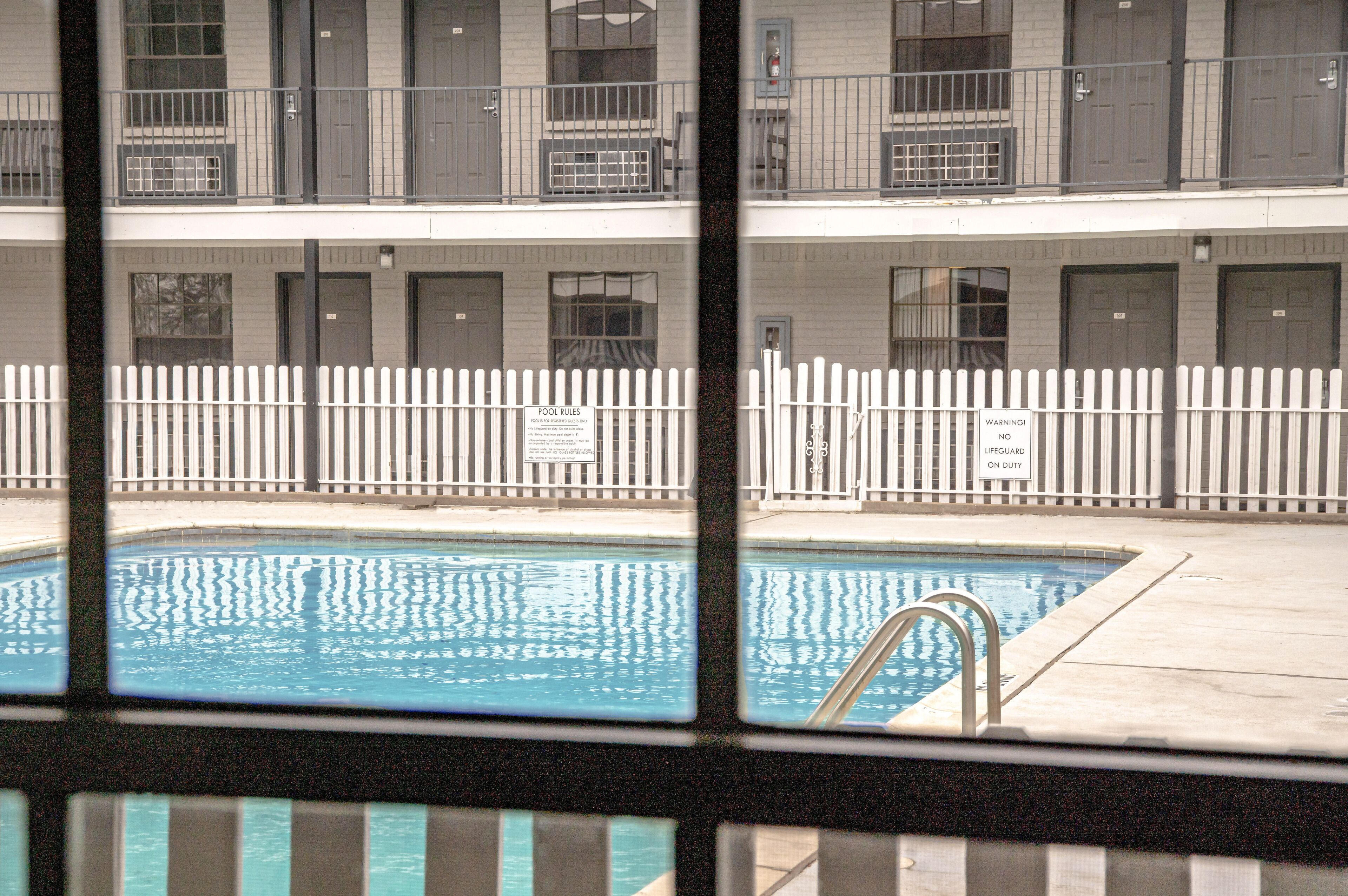 A pool area with a white fence, a lifeguard sign, and a building with multiple doors and windows in the background.