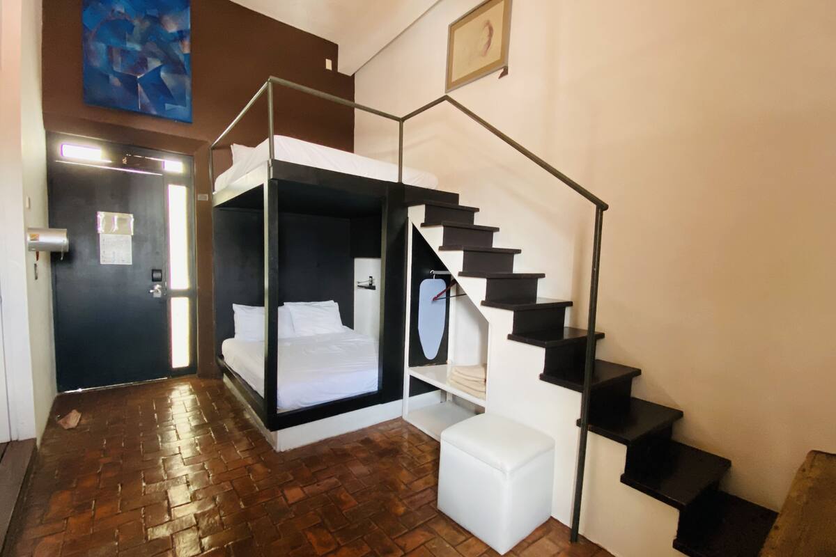Una habitación con una cama individual doble, un pequeño taburete blanco y una escalera con barandillas negras.
