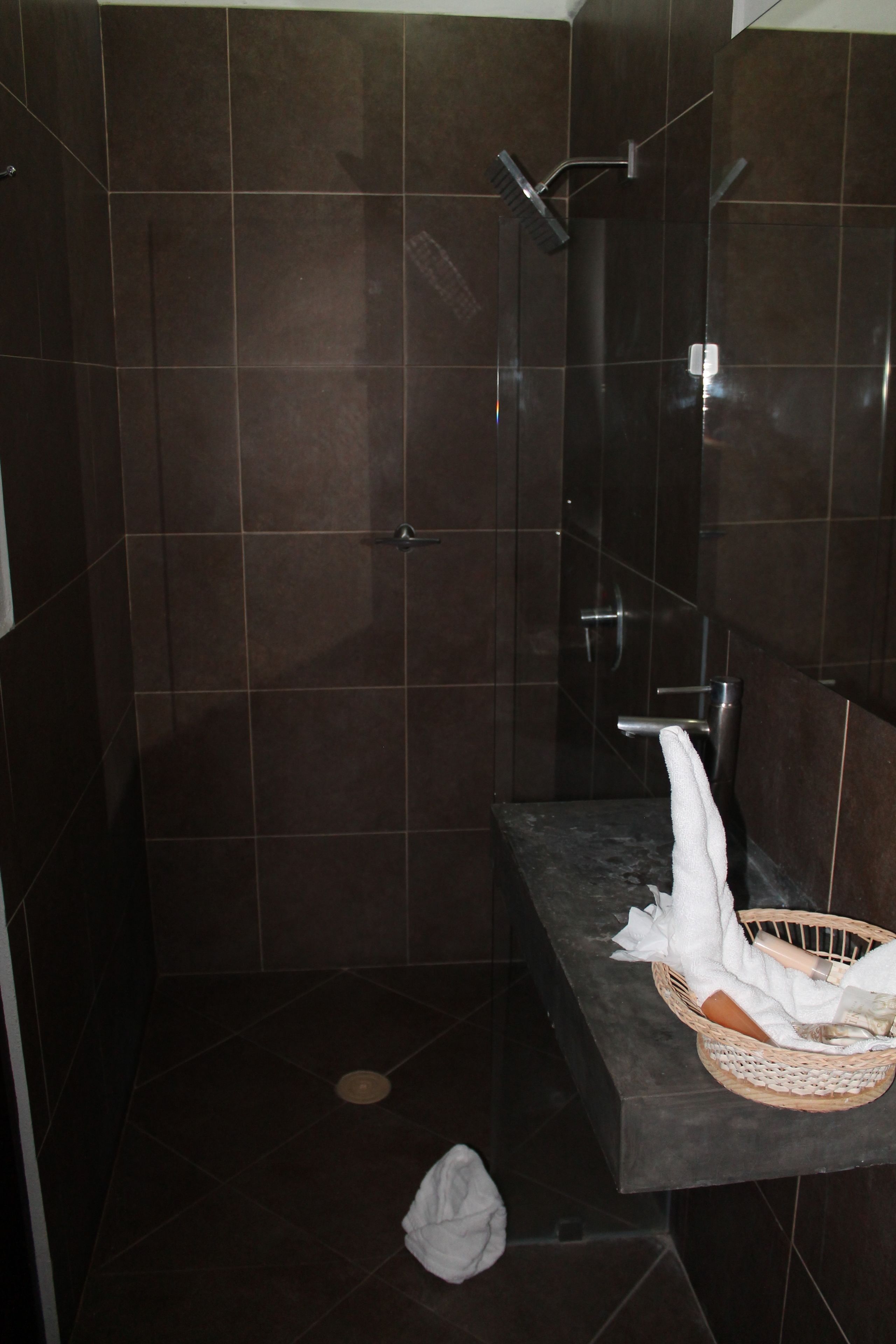 Un baño con baldosas oscuras, una ducha con cabezal de ducha manual, un perchero y una cesta sobre un banco.