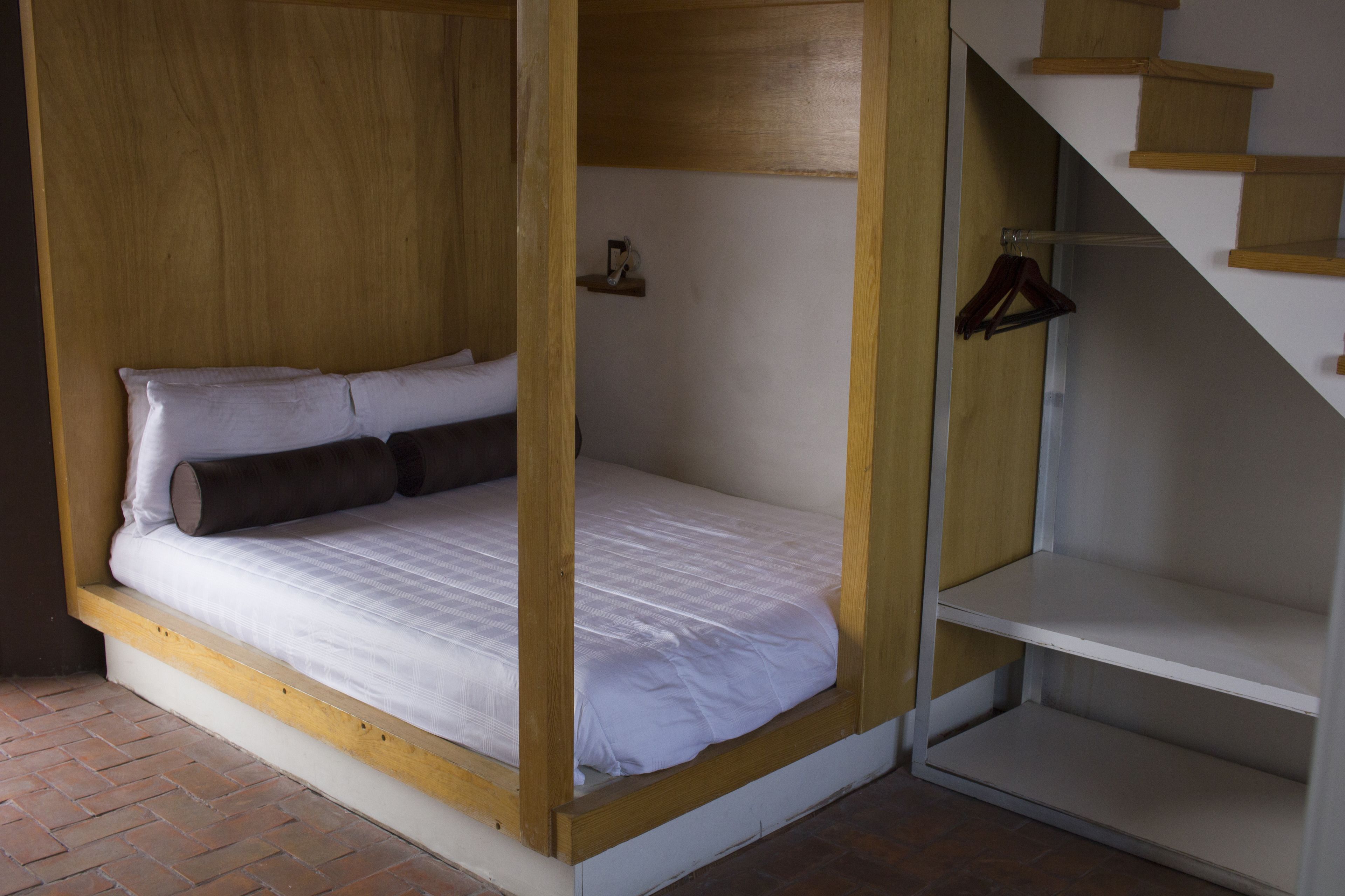 A single bed with a wooden frame and white bedding, a small shelf with a cup and a hanger, and a staircase with wooden steps.