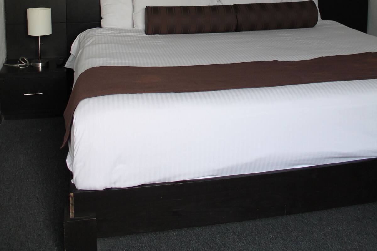 Una cama bien tendida con ropa de cama blanca y marrón, un cabecero oscuro y un cuadro en la pared.