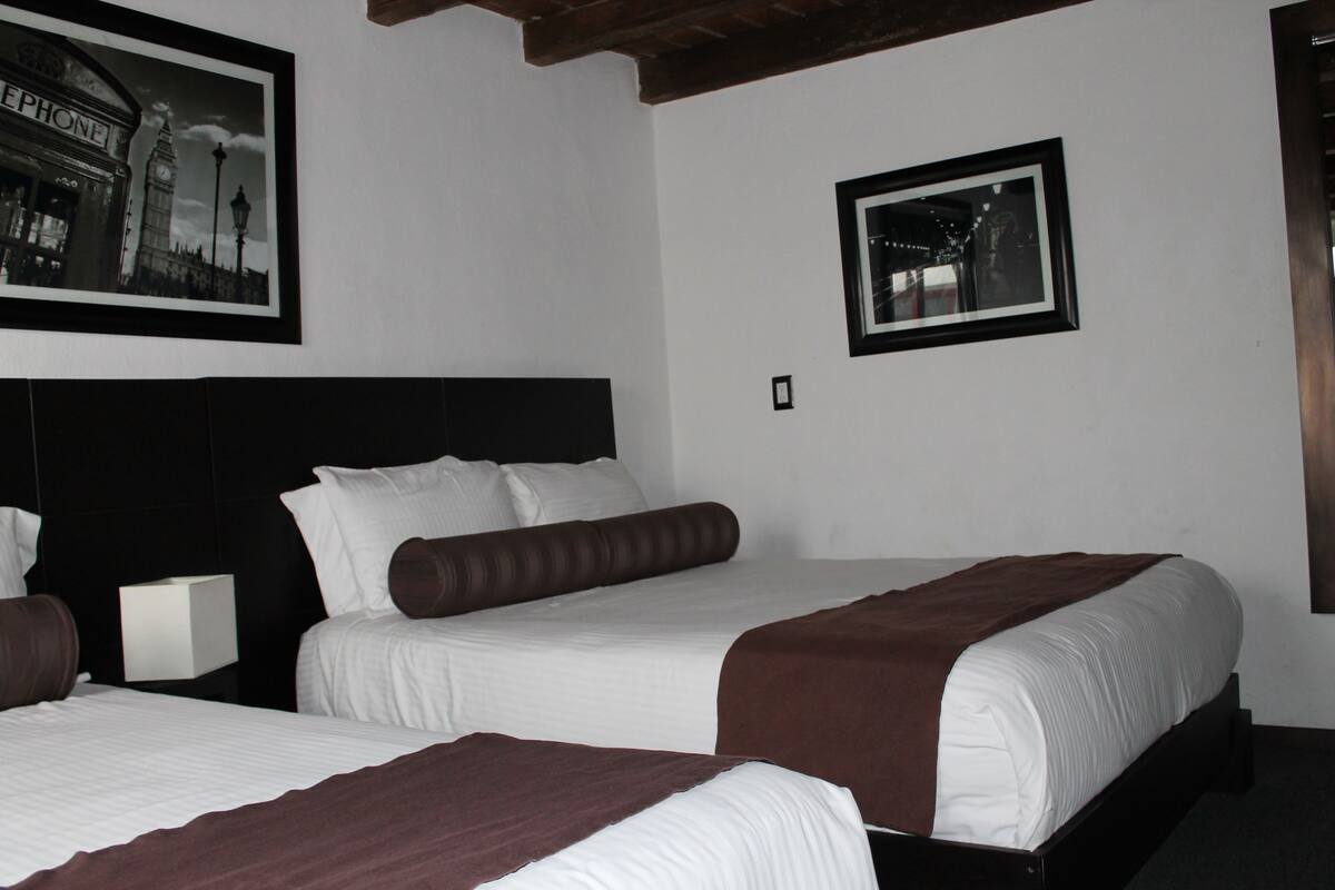 Habitación de hotel con dos camas, cabecero negro, ropa de cama blanca y dos cuadros en las paredes.