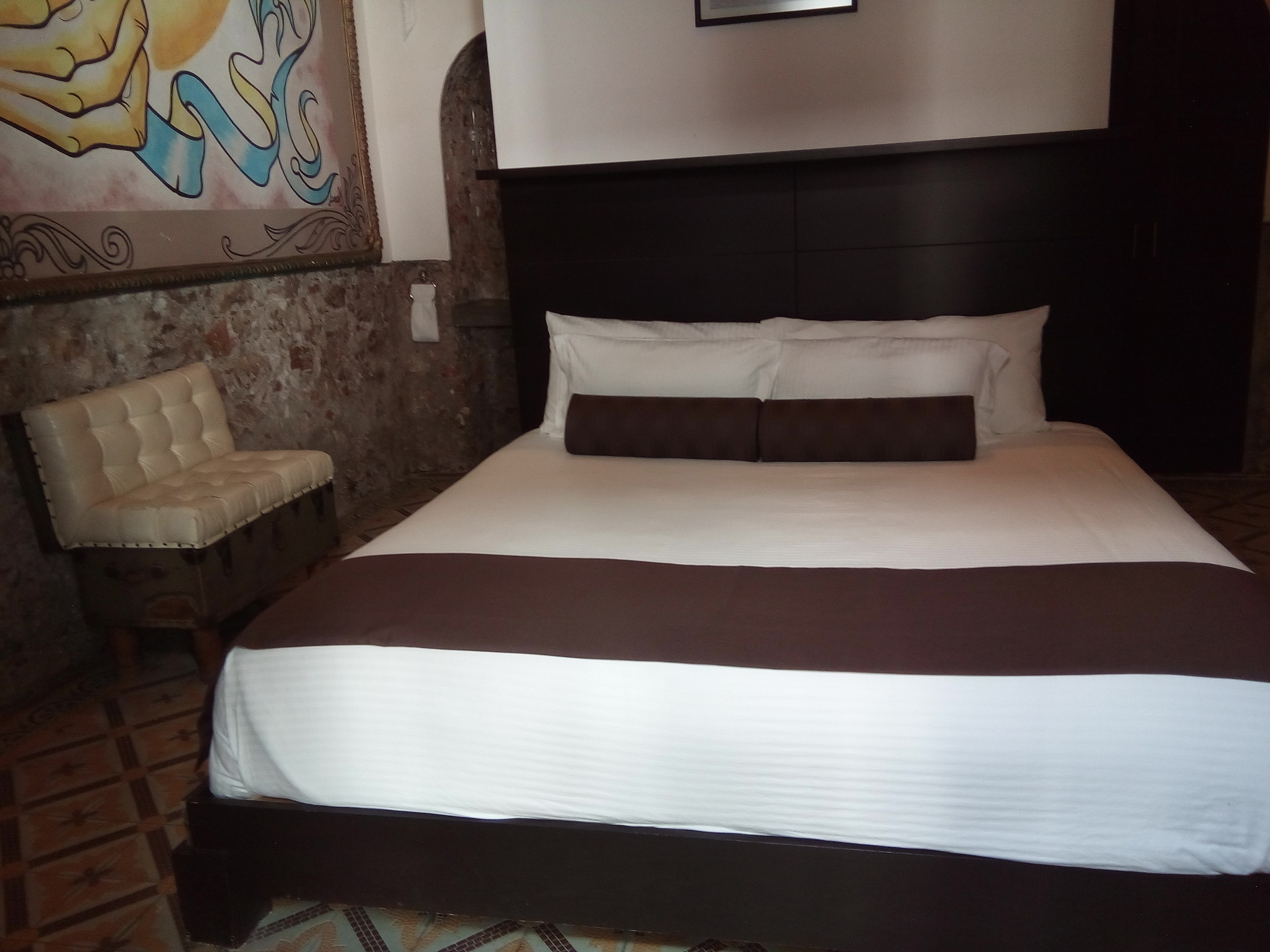 Une chambre d’hôtel avec un grand lit, une tête de lit, une chaise et un mur orné d’un tableau.