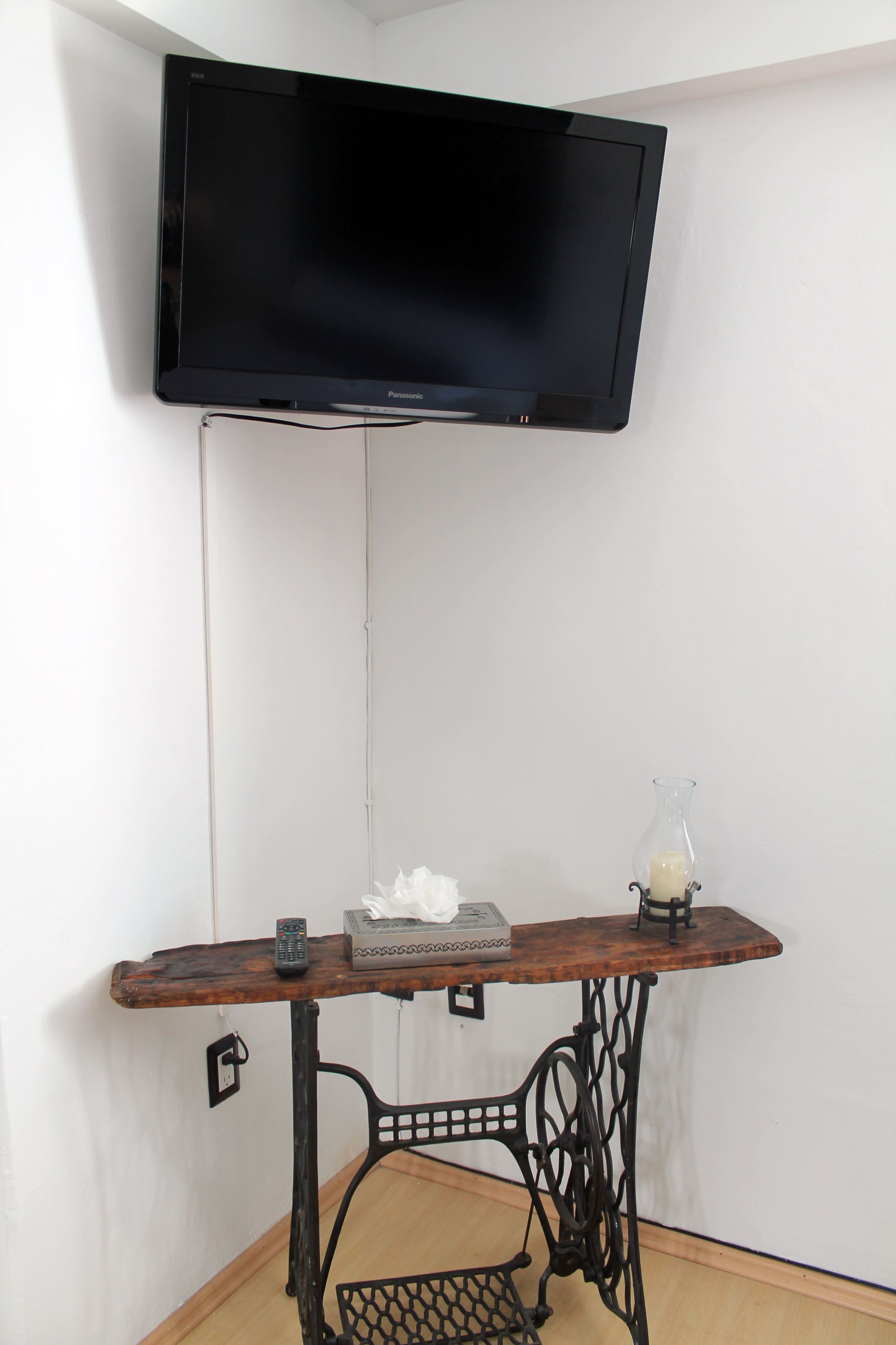 A corner room with a flat-screen TV mounted on the wall, a wooden table with a sewing machine, a remote control, and a small vase with a candle.